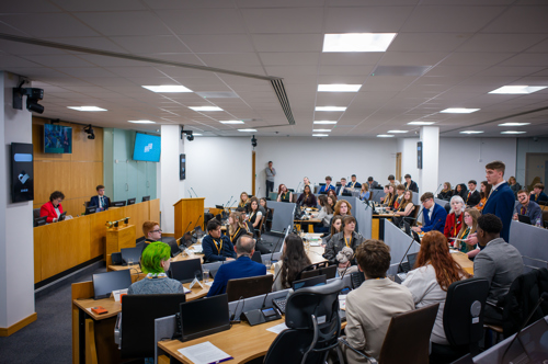 Mae un aelod o WYP yn sefyll ac yn cyflwyno araith tra bod yr aelodau eraill yn gwrando - yn y siambr