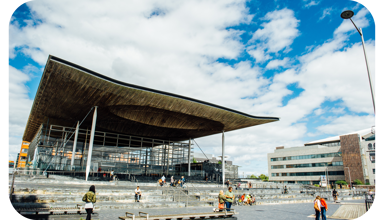 Ffotograff o du blaen adeilad y Senedd ym Mae Caerdydd. Mae'r adeilad wedi'i wneud o frics llwyd, ac mae rhai trawstiau gwyn i'w gweld. Mae arwydd sy'n dweud 'Senedd Cymru'. Yn y blaendir, mae cerddwyr a beicwyr yn mynd heibio. Mae rhai pobl yn eistedd ar y grisiau y tu allan i adeilad y Senedd.