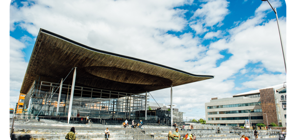 Ffotograff o du blaen adeilad y Senedd ym Mae Caerdydd. Mae'r adeilad wedi'i wneud o frics llwyd, ac mae rhai trawstiau gwyn i'w gweld. Mae arwydd sy'n dweud 'Senedd Cymru'. Yn y blaendir, mae cerddwyr a beicwyr yn mynd heibio. Mae rhai pobl yn eistedd ar y grisiau y tu allan i adeilad y Senedd.