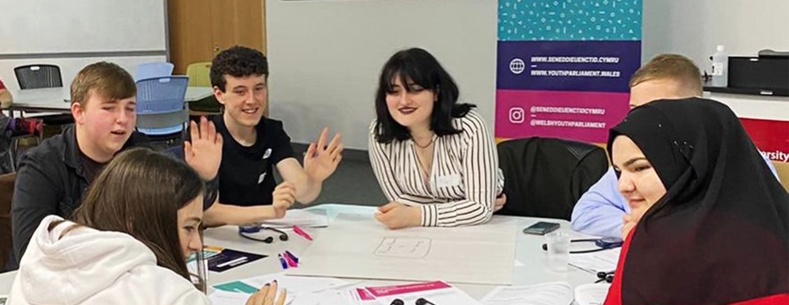 Grŵp o bobl ifanc yn eistedd wrth fwrdd yn siarad â'i gilydd.