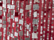 Mae rhesi o ddarnau ffabrig sgwâr bach mewn gwyn, llwyd a choch yn hongian mewn llinynnau fertigol, gan greu effaith llen haenog yn erbyn cefndir coch.