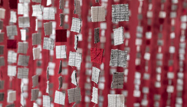 Mae rhesi o ddarnau ffabrig sgwâr bach mewn gwyn, llwyd a choch yn hongian mewn llinynnau fertigol, gan greu effaith llen haenog yn erbyn cefndir coch.